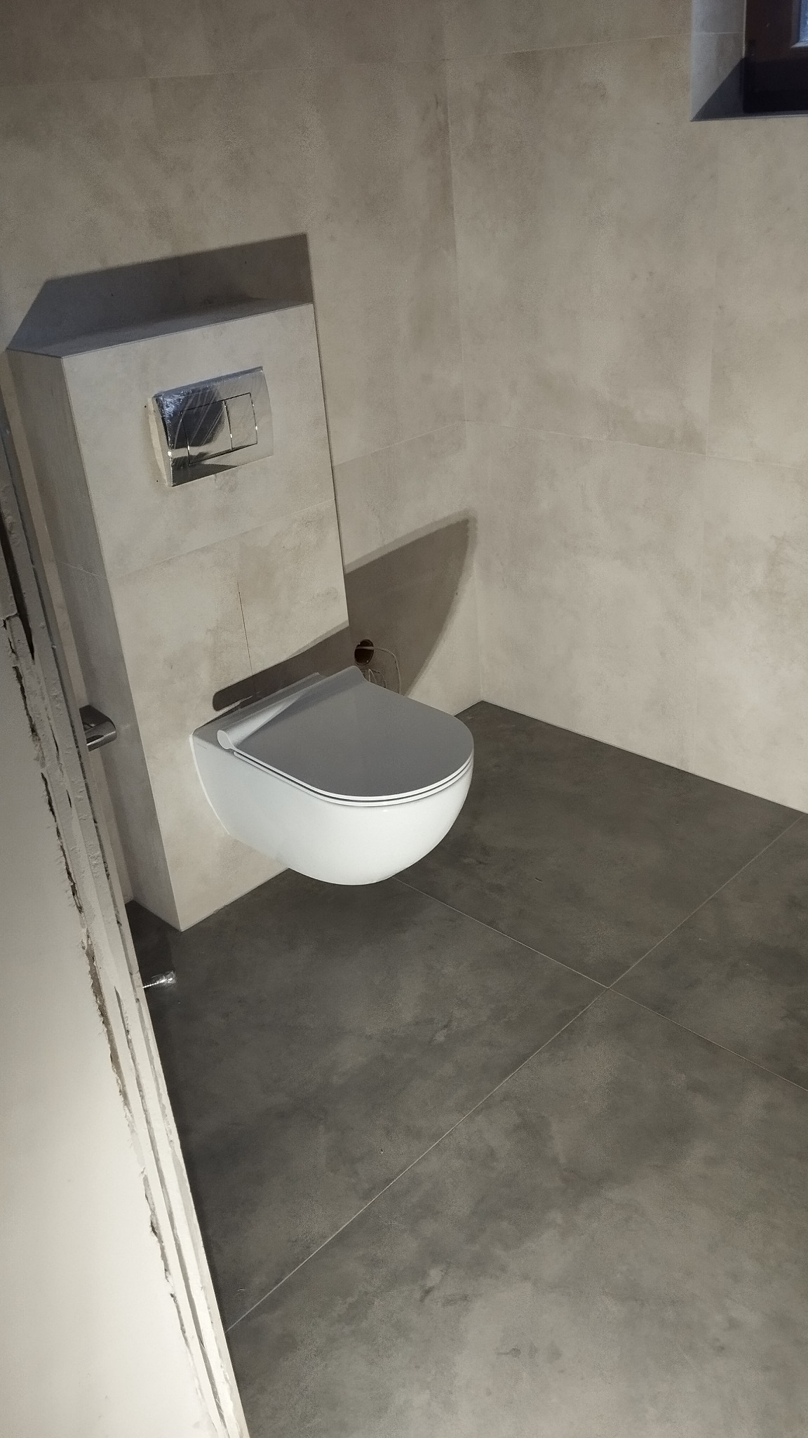 Nowoczesna, podwieszana toaleta z chromowanym przyciskiem spłukiwania, zamontowana na ścianie wyłożonej płytkami imitującymi beton, z szarymi płytkami podłogowymi.
