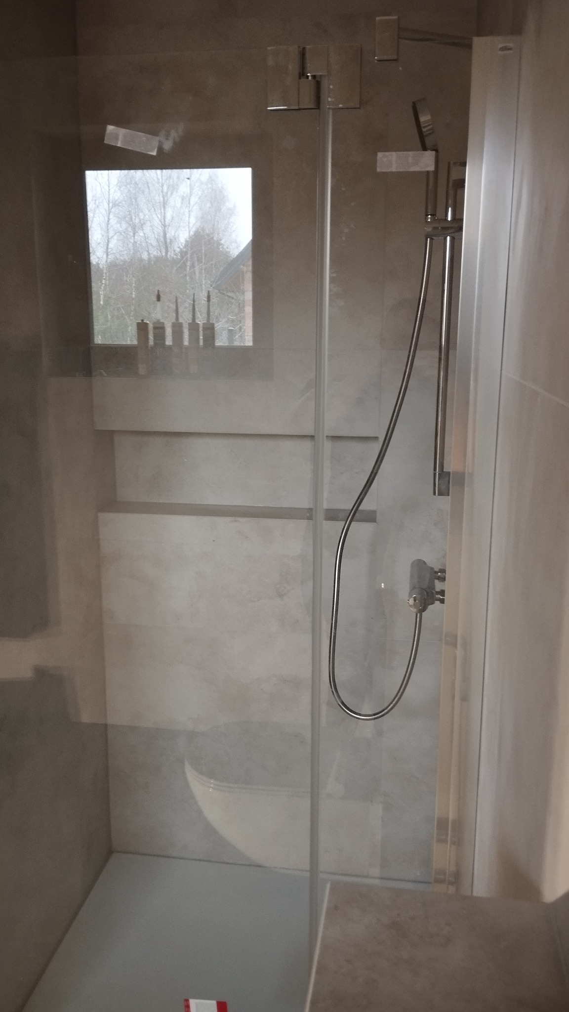 Nowoczesna kabina prysznicowa z deszczownicą i widokiem na zewnątrz przez małe okno, neutralna kolorystyka płytek.