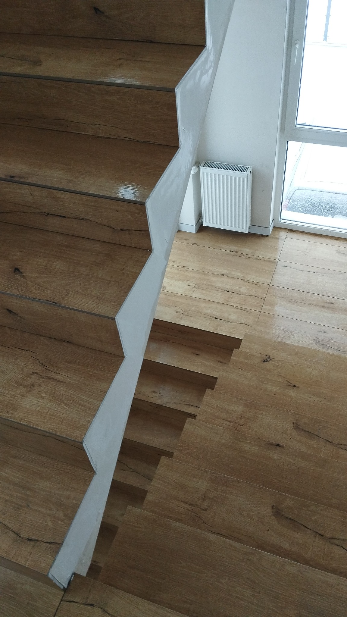 Schody drewniane z białymi wspornikami, dopasowane do podłogi z paneli o wzorze drewna, w tle biały grzejnik i okno.