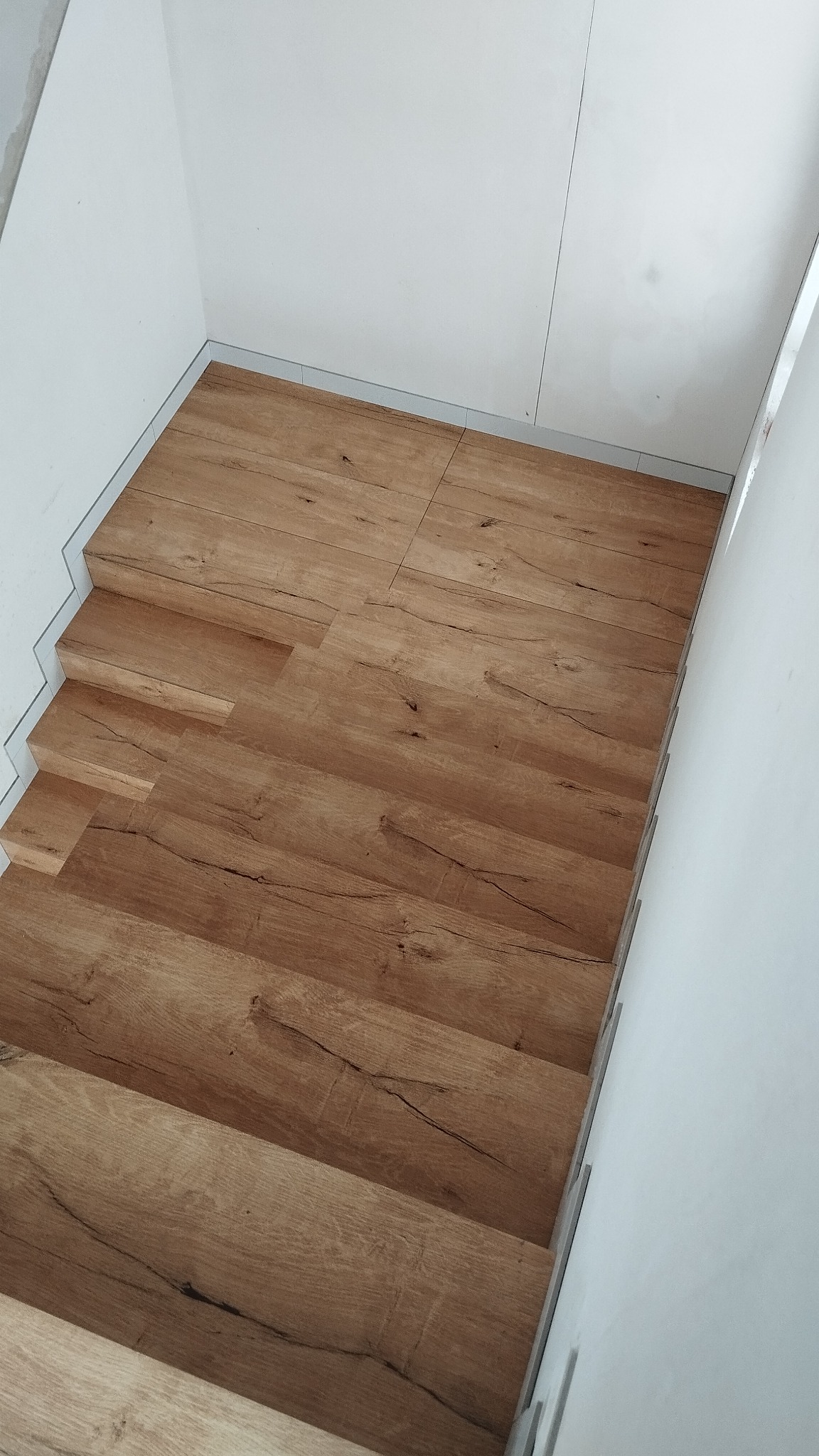 Schody wyłożone panelami imitującymi drewno, prowadzące w dół, z widocznymi detalami struktury drewna i białymi ścianami po bokach.