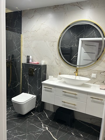 Elegancka łazienka z czarno-białymi marmurkowymi płytkami, wiszącą toaletą, okrągłym lustrem z podświetleniem LED i złotymi akcentami.