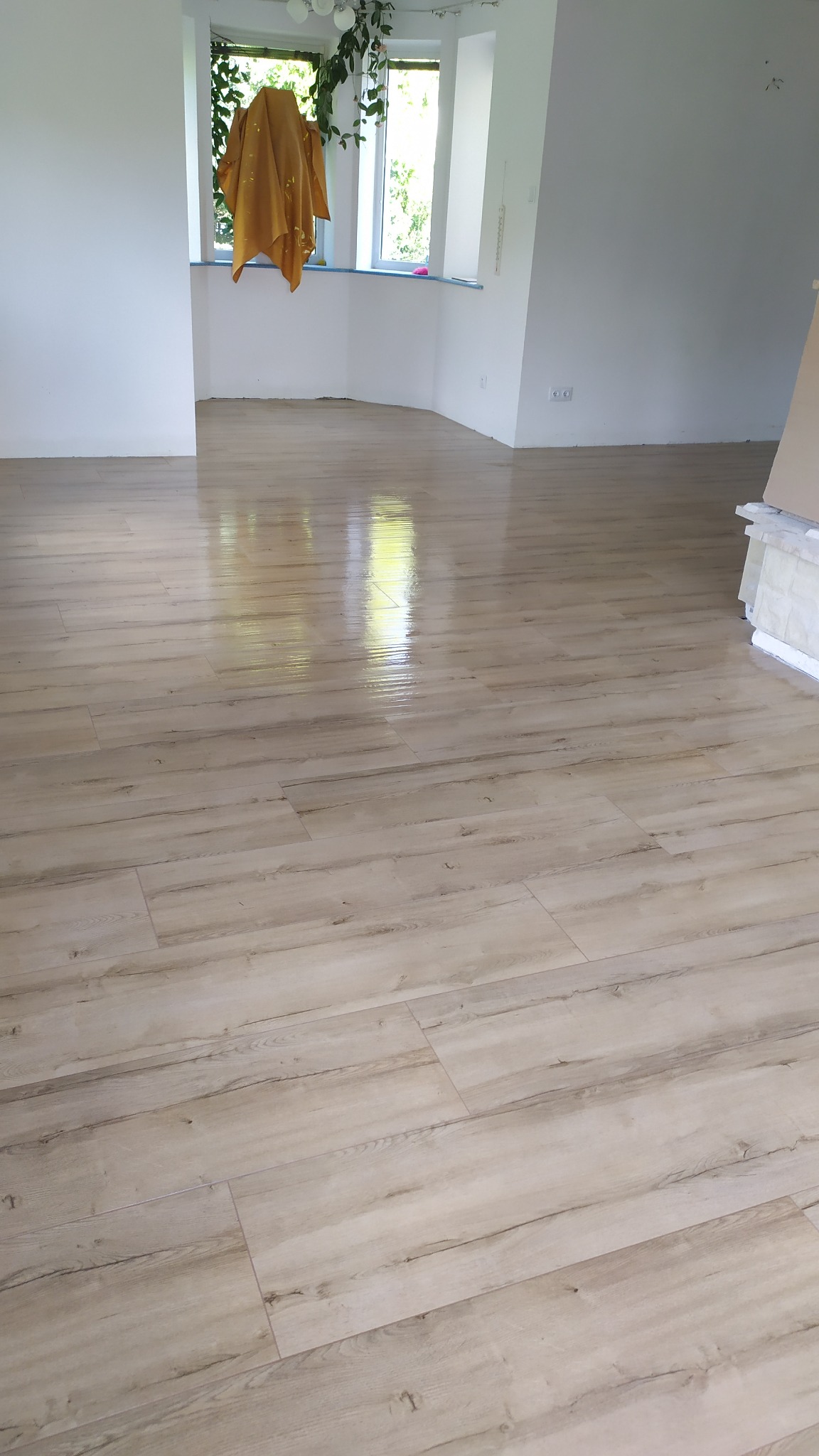 Jasne panele podłogowe imitujące drewno, ułożone w przestronnym, słonecznym pokoju z dużym oknem i stertą płytek przy ścianie.