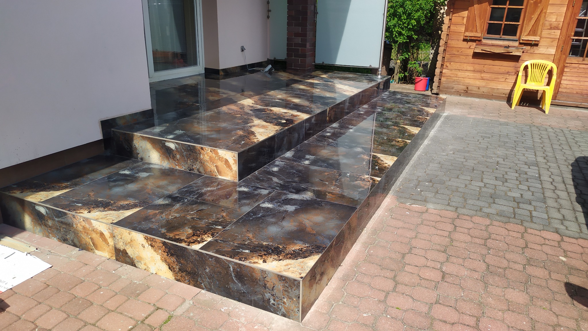 Schody wejściowe wyłożone błyszczącymi, wielkoformatowymi płytkami imitującymi marmur w odcieniach brązu i szarości, prowadzące do budynku z jasną elewacją i drewnianą chatką obok, na kostce brukowej.