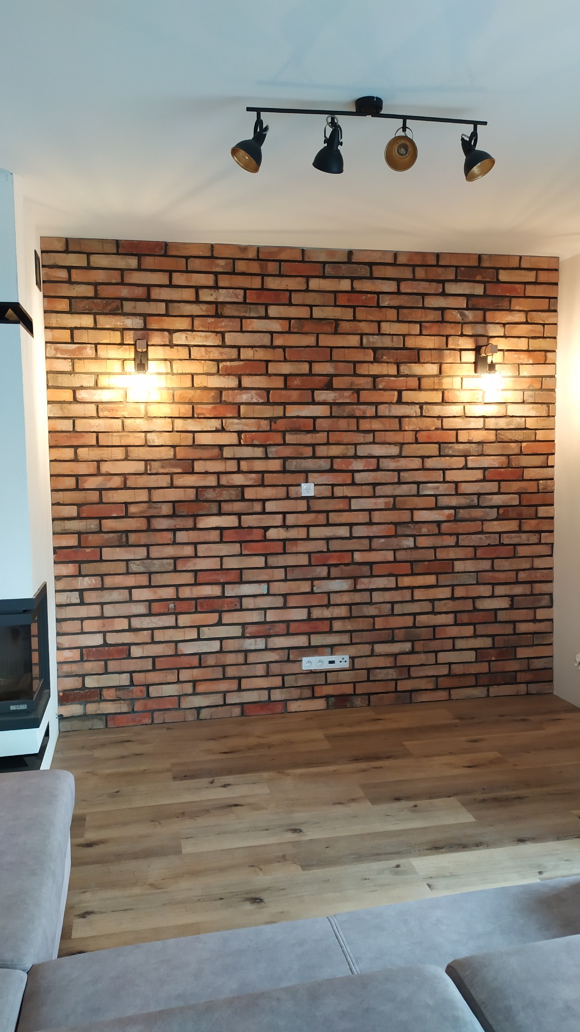 Ściana wykończona cegłą klinkierową z zamontowanymi kinkietami, oświetlenie sufitowe na szynie, panele podłogowe, fragment sofy w salonie.