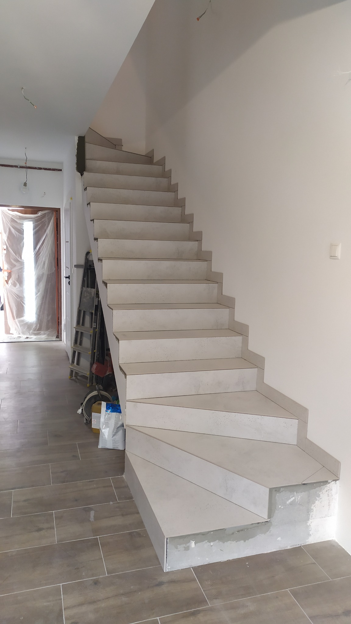 Wykończone schody z szarych płytek w surowym wnętrzu, widoczna drabina i materiały budowlane, podłoga wyłożona panelami imitującymi drewno.