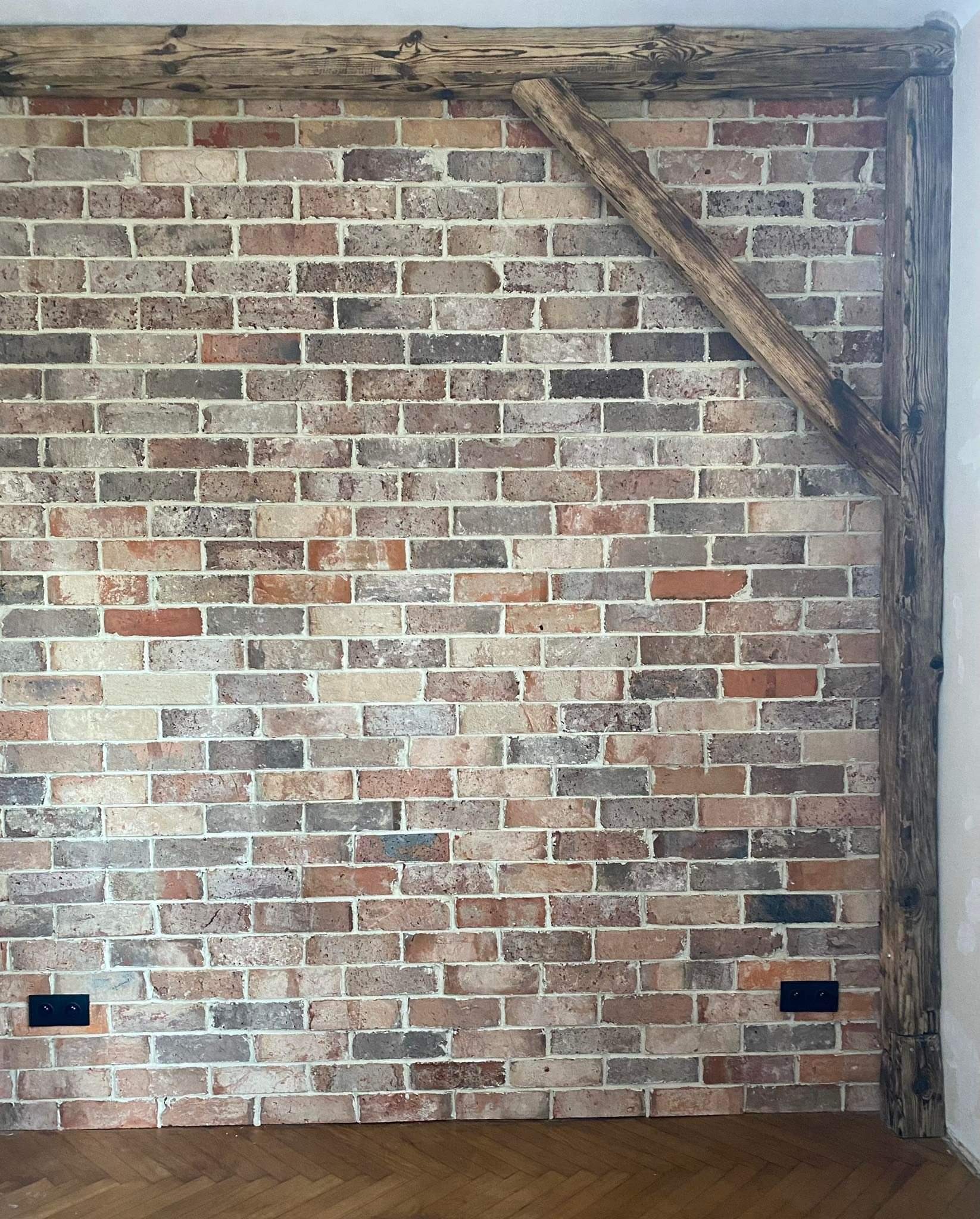 Ściana z cegły klinkierowej w odcieniach brązu, beżu i szarości, z drewnianą konstrukcją belki w rogu i nad nią, widoczne gniazdka elektryczne w kolorze czarnym na ścianie.