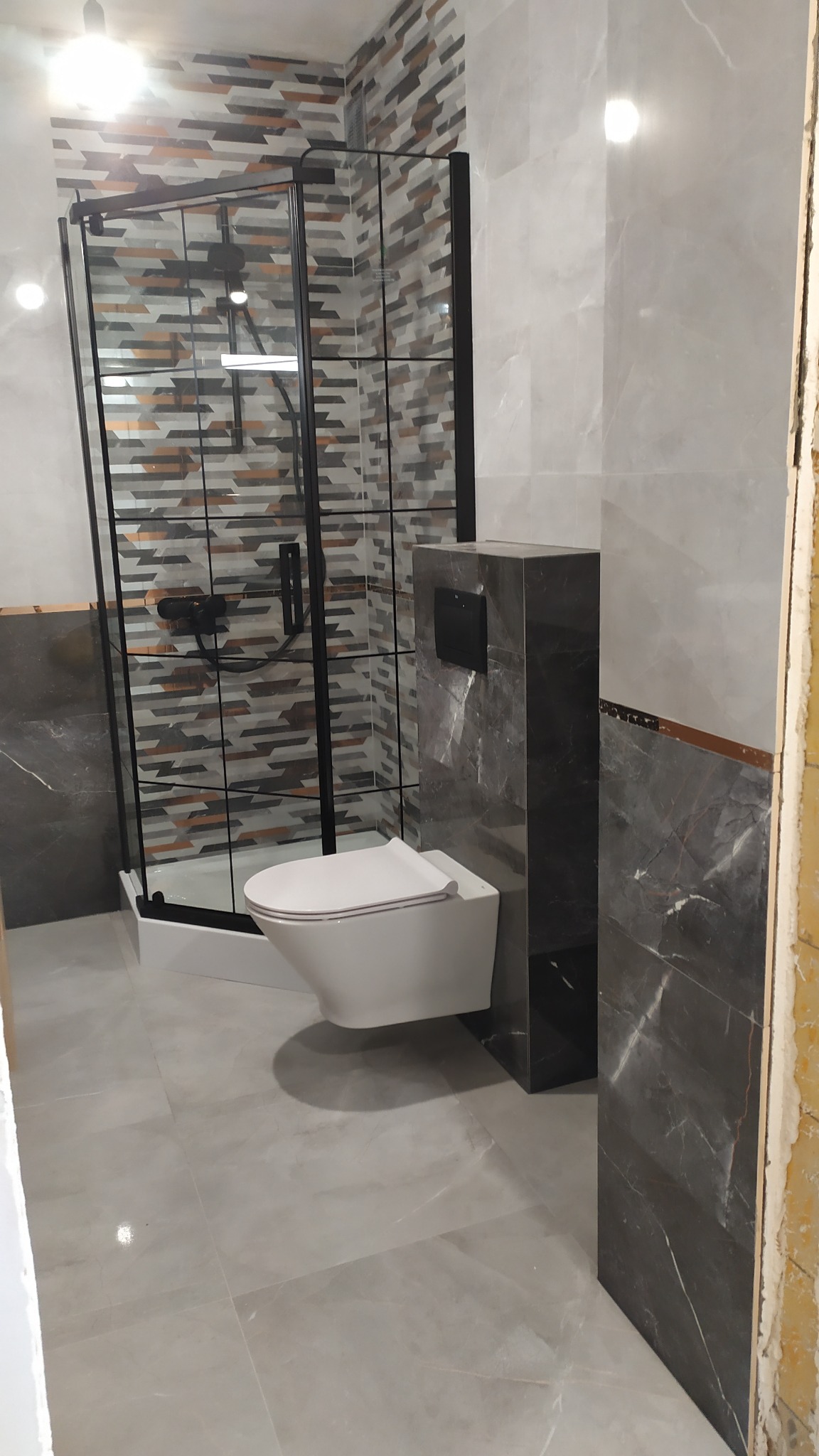 Nowoczesna łazienka z kabiną prysznicową z czarnymi profilami, podwieszaną toaletą i szarą marmurową okładziną ścienną z dekoracyjnym pasem. Widoczne płytki podłogowe w odcieniach szarości.