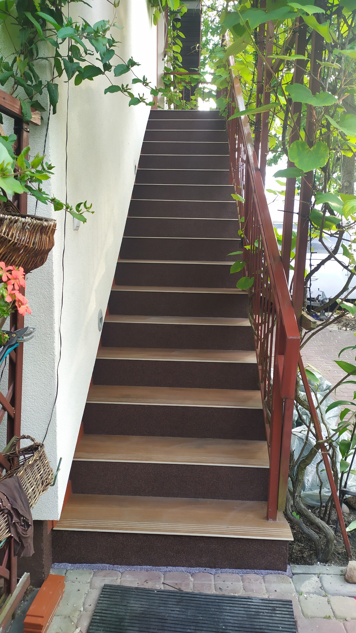 Zewnętrzne schody obłożone brązową, ziarnistą okładziną z jasnymi paskami antypoślizgowymi, prowadzące do góry, otoczone zielenią i czerwoną metalową barierką.
