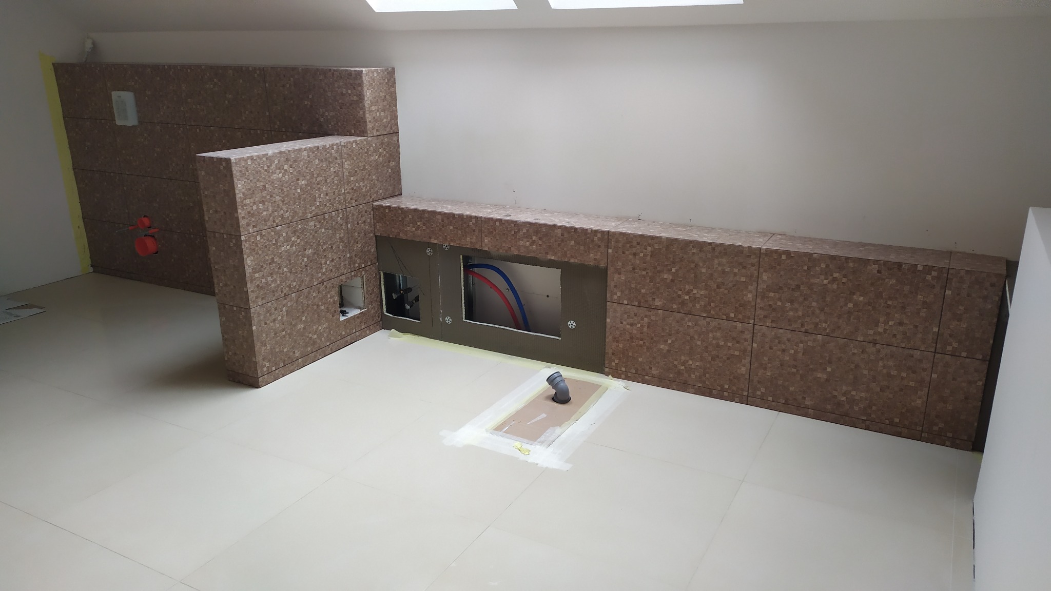 Łazienka w trakcie wykańczania, widoczne płytki mozaikowe na ścianach i zabudowie, przygotowane otwory na instalacje, podłoga wyłożona jasnymi płytkami, sufit ze świetlikami.