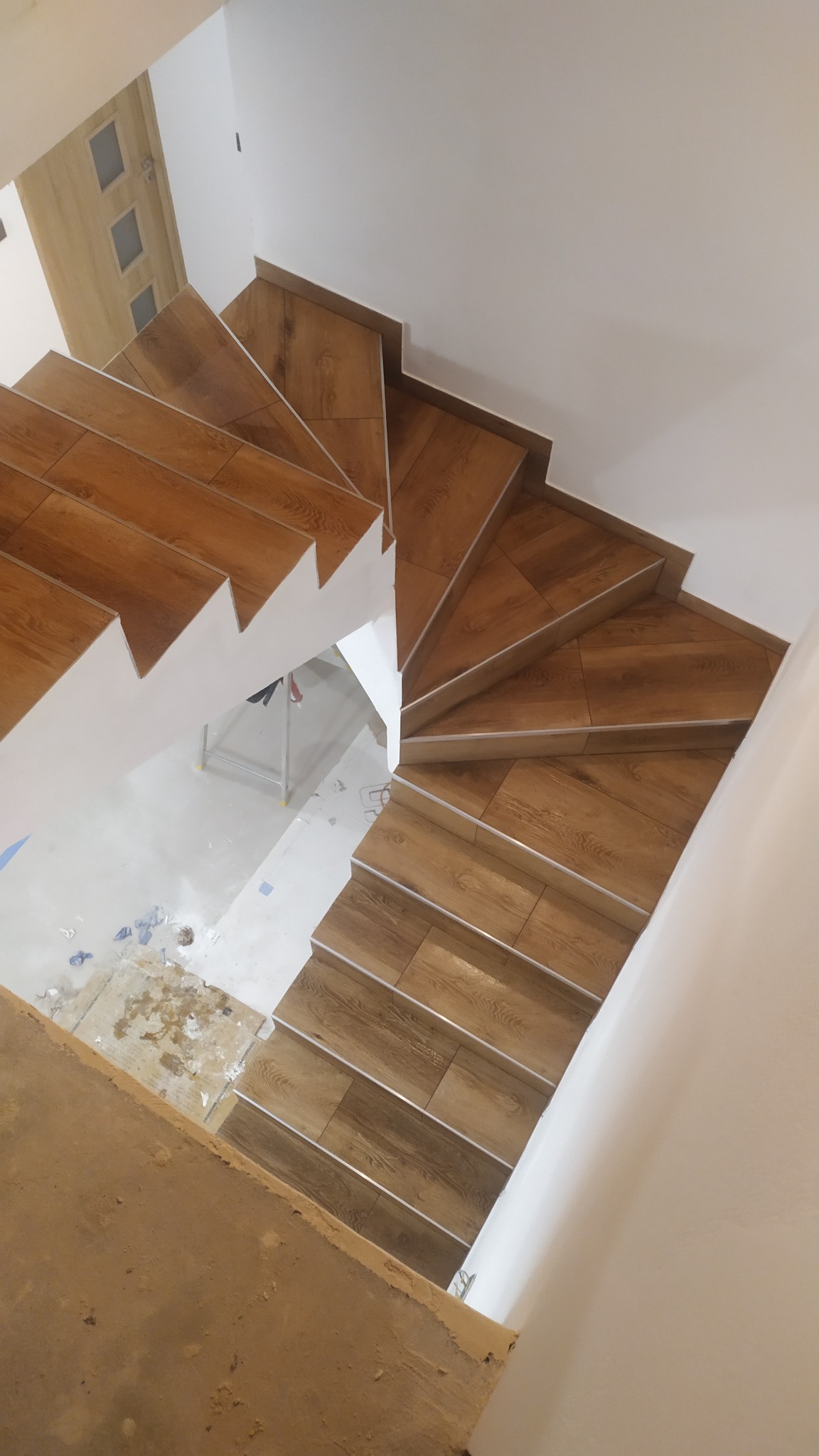 Wewnętrzne spiralne schody wyłożone płytkami imitującymi drewno, z aluminiowymi listwami na krawędziach stopni, widok z góry na etapie prac wykończeniowych z widocznymi zabrudzeniami na podłodze...