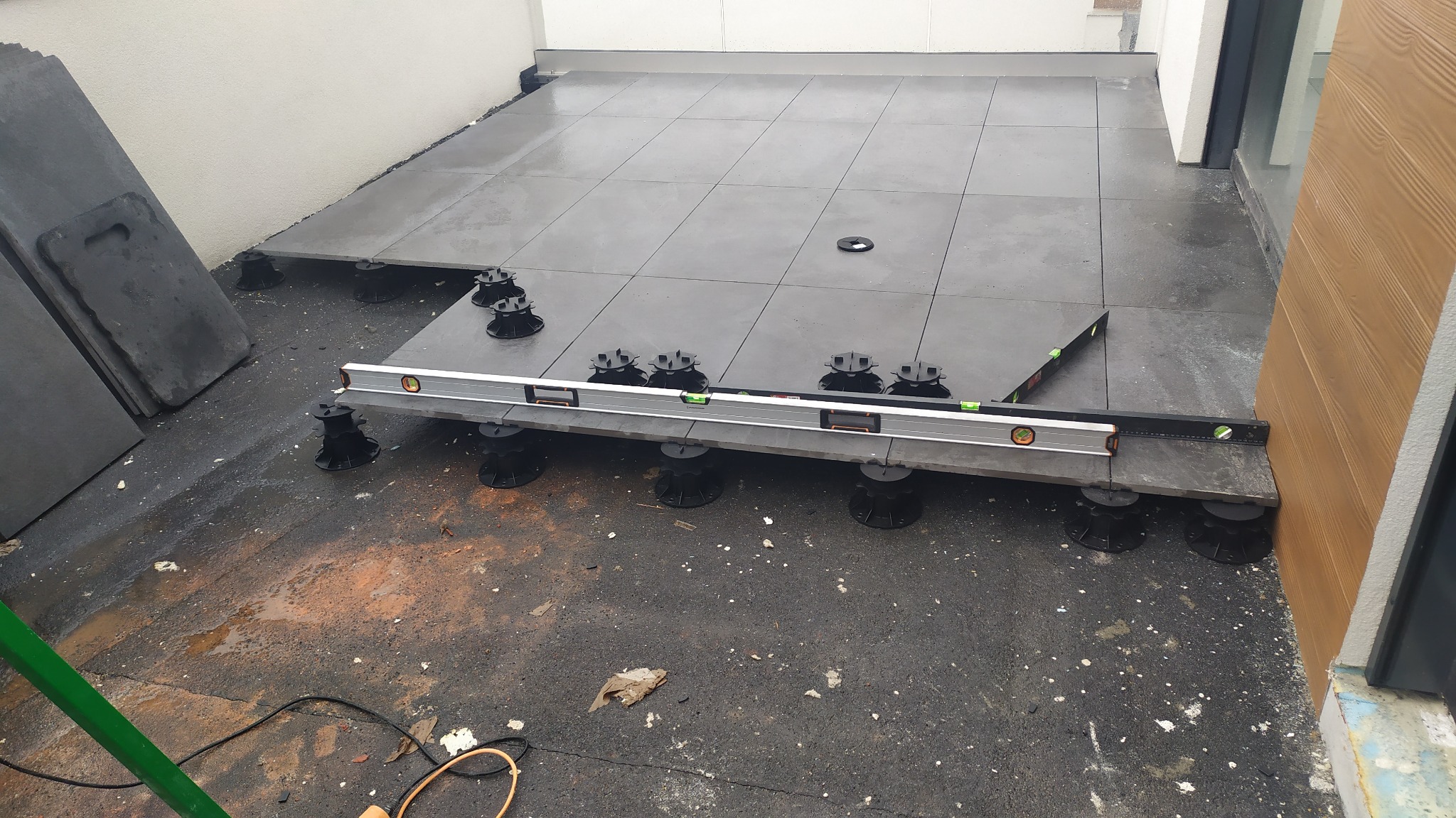 Montaż ciemnoszarych płyt tarasowych na regulowanych wspornikach, poziomowanie za pomocą poziomicy, widoczne płyty czekające na ułożenie i fragment elewacji.