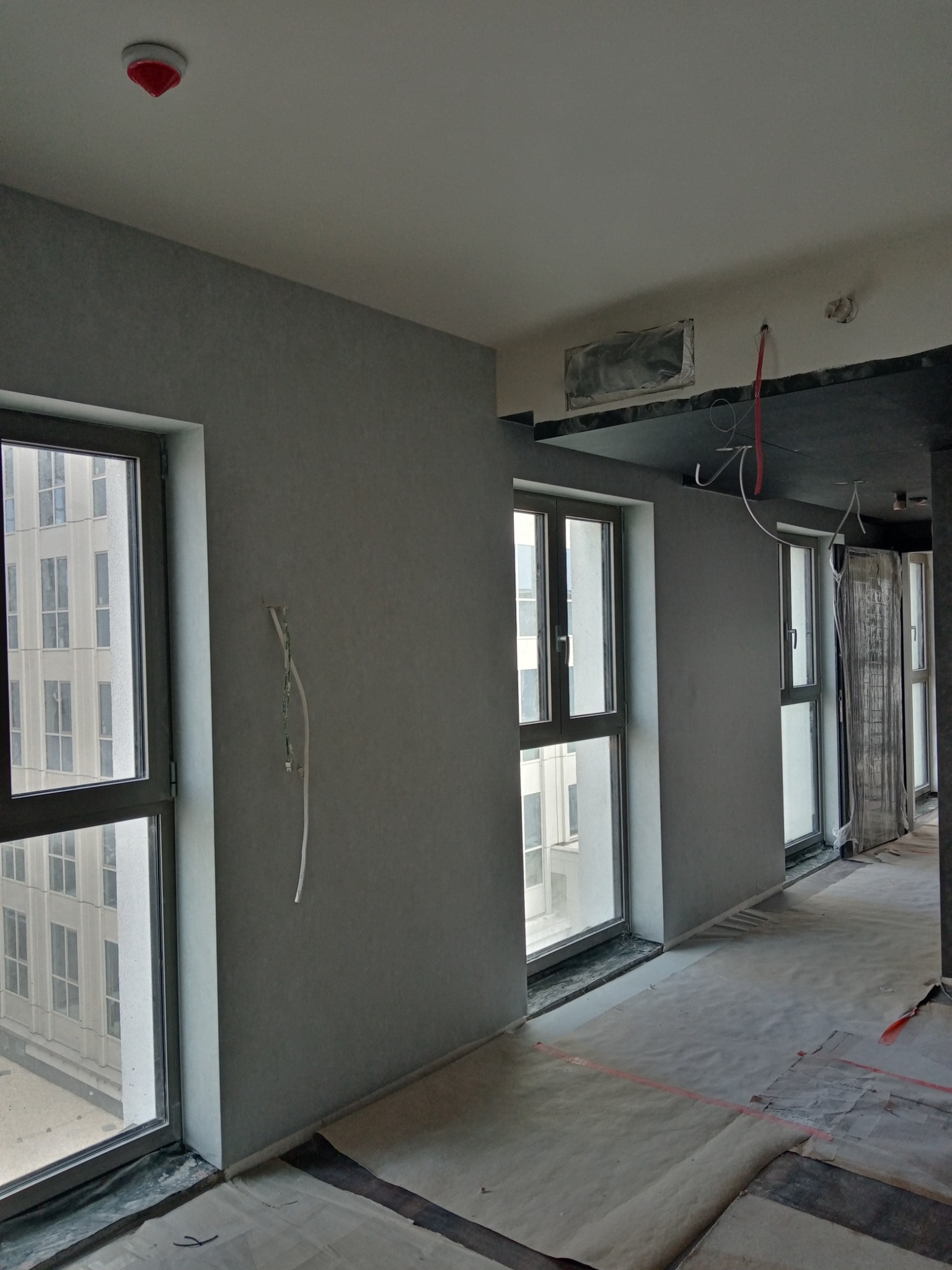 Wnętrze w trakcie prac wykończeniowych: szare ściany, okna z widokiem na budynek, zabezpieczona podłoga, wystające kable elektryczne i częściowo podwieszony sufit.