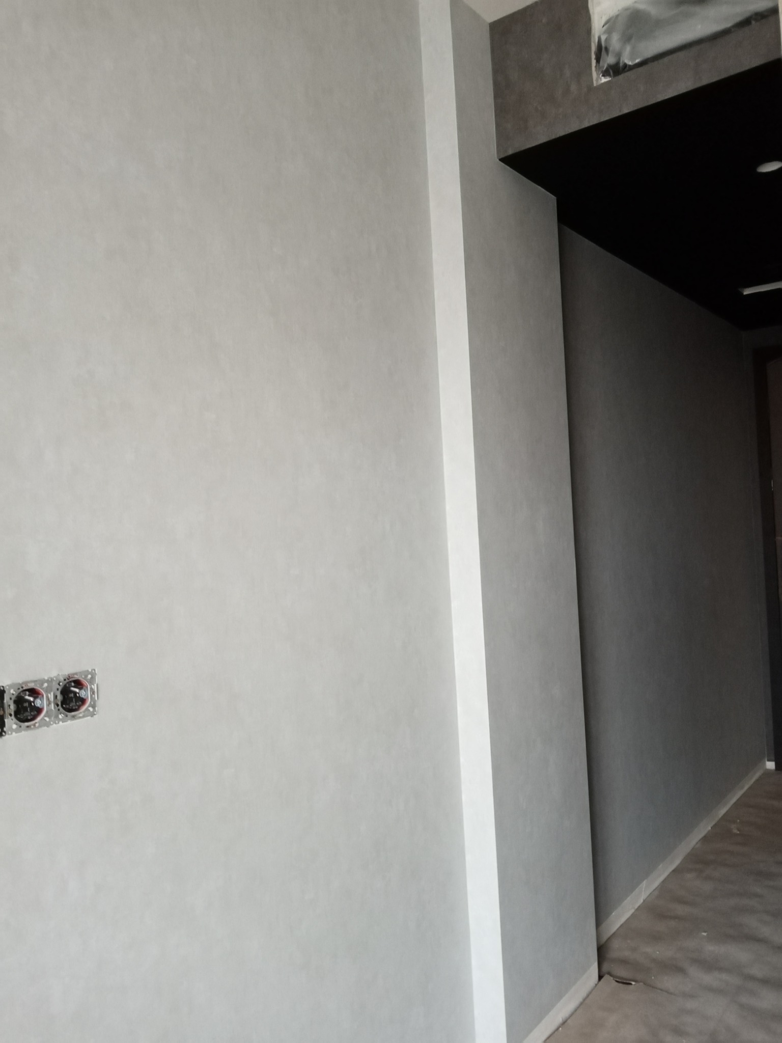 Świeżo pomalowana ściana w odcieniach szarości z widocznymi puszkami elektrycznymi przed montażem gniazdek, fragment sufitu podwieszanego z czarną wnęką.