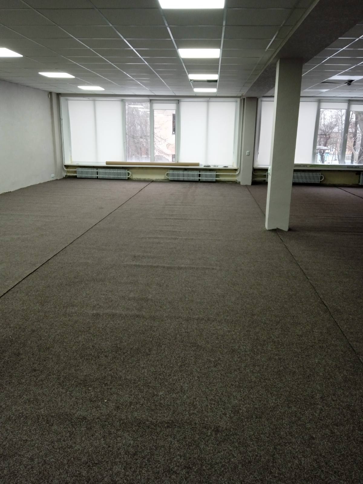 Duże, puste pomieszczenie biurowe z nową, ciemnoszarą wykładziną dywanową, widoczne grzejniki pod oknami z roletami i kolumna nośna w tle.