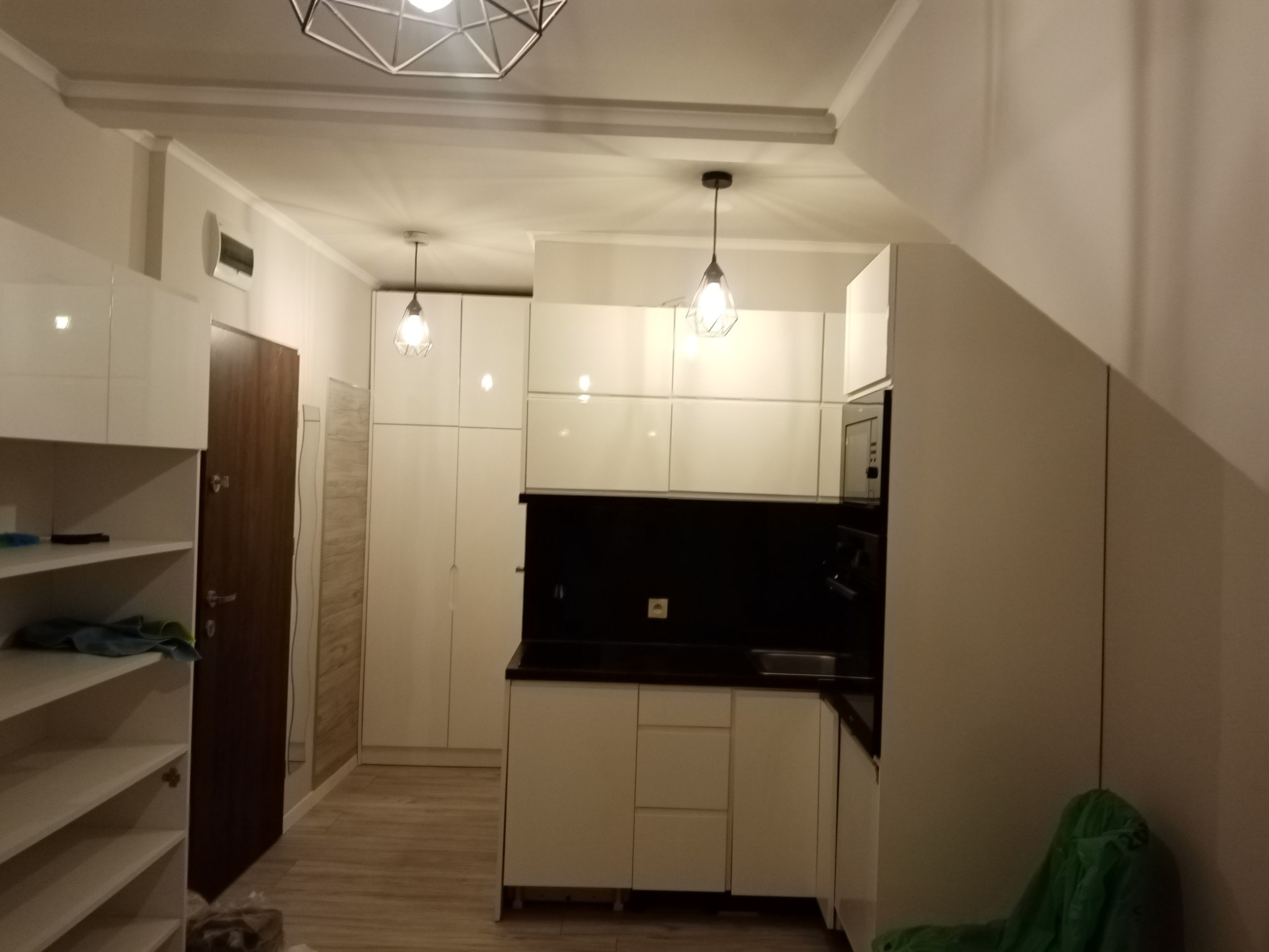 Nowoczesna kuchnia w odcieniach bieli z czarnym blatem, oświetlona wiszącymi lampami z geometrycznym kloszem, widok na regał z półkami i fragment podłogi z panelami.