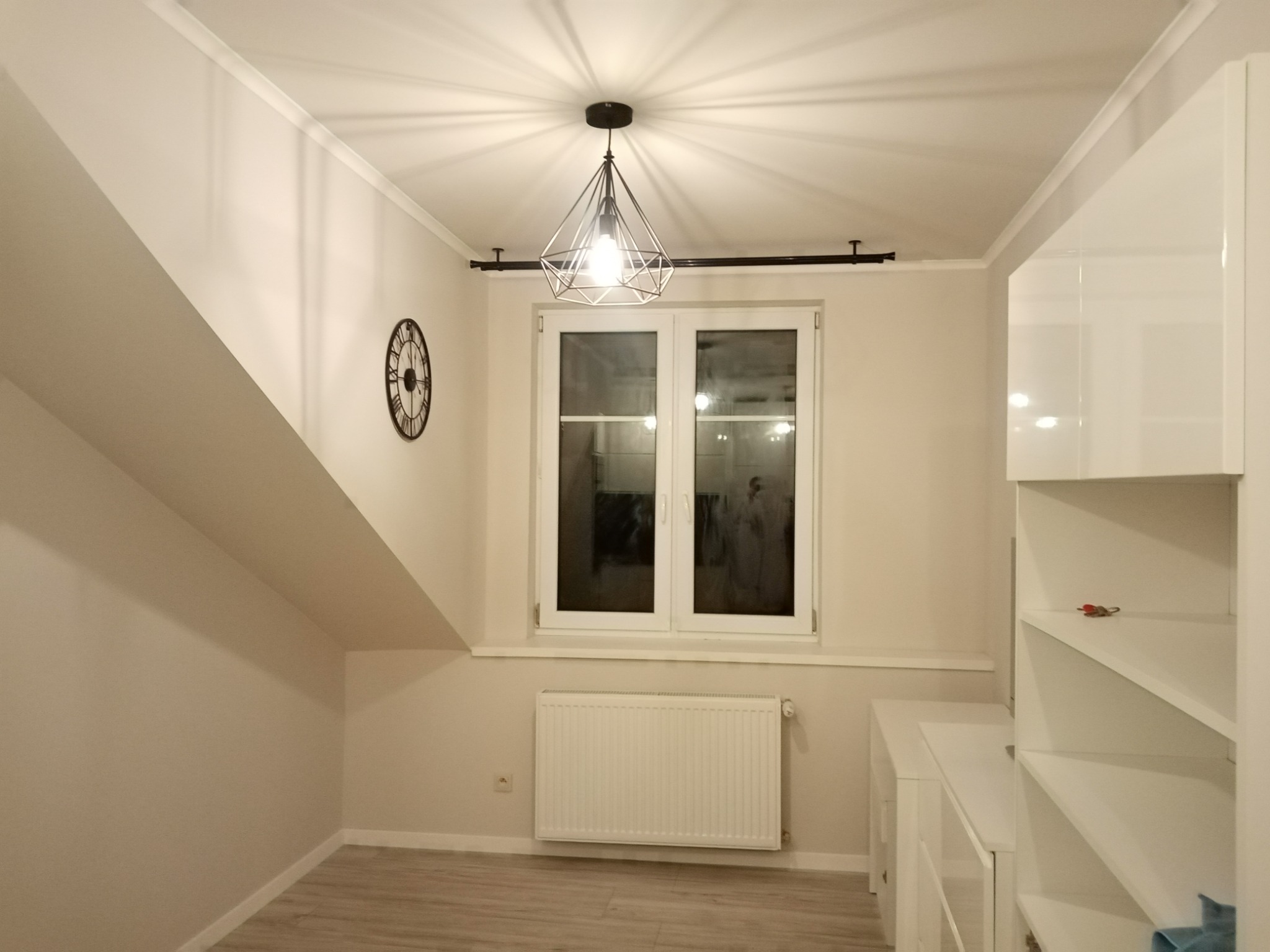 Jasne wnętrze pokoju na poddaszu z oknem, grzejnikiem, białą zabudową meblową i lampą w stylu industrialnym, widoczne wykończenia pod skosem.