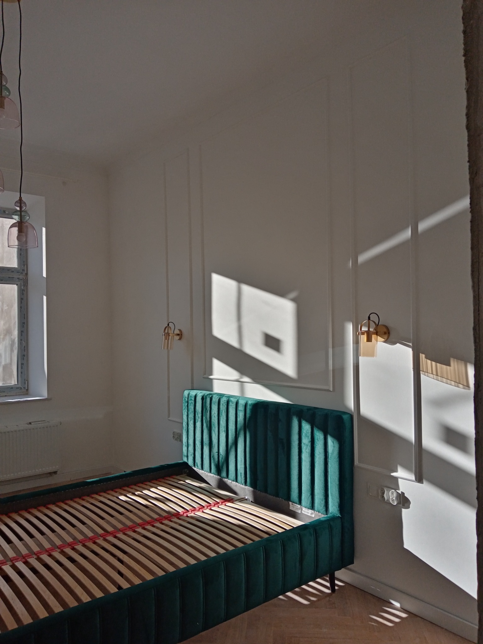 Pusta rama łóżka z zielonym, welurowym zagłówkiem i bokami w pokoju z białymi ścianami z dekoracyjnymi listwami i oświetleniem ściennym, oświetlonym promieniami słońca.