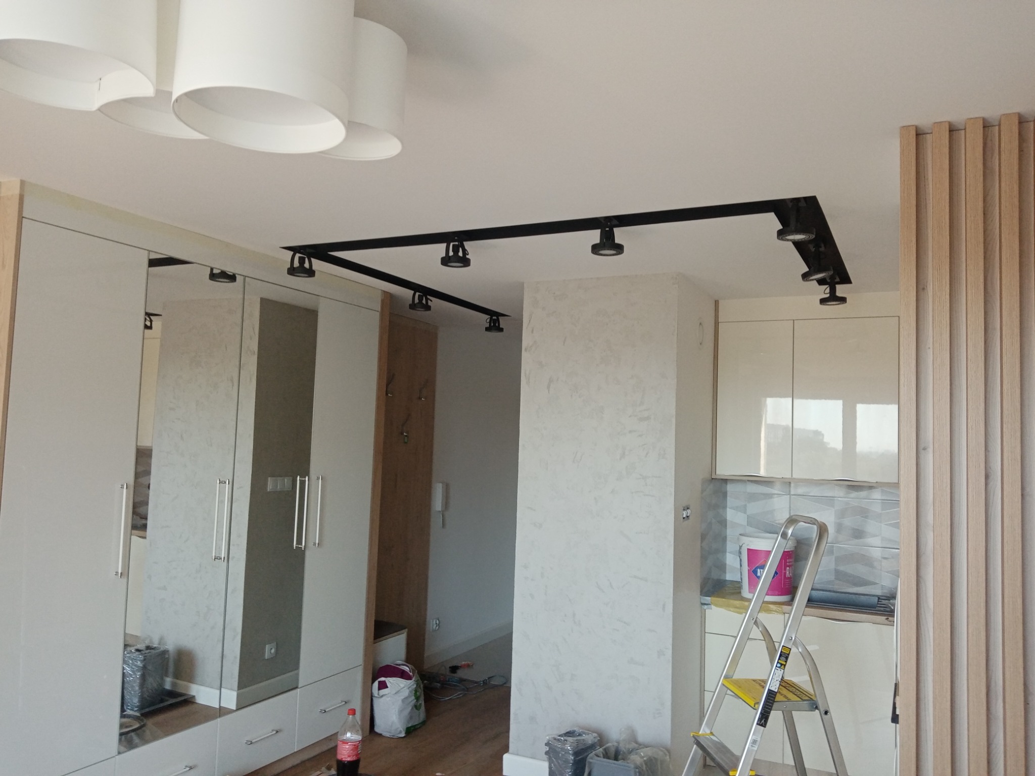 Wnętrze mieszkania w Krakowie w trakcie prac wykończeniowych: zamontowane oświetlenie szynowe na suficie, widoczny fragment zabudowy szafy z lustrami, drabina oparta o ścianę w kuchni, puszka...