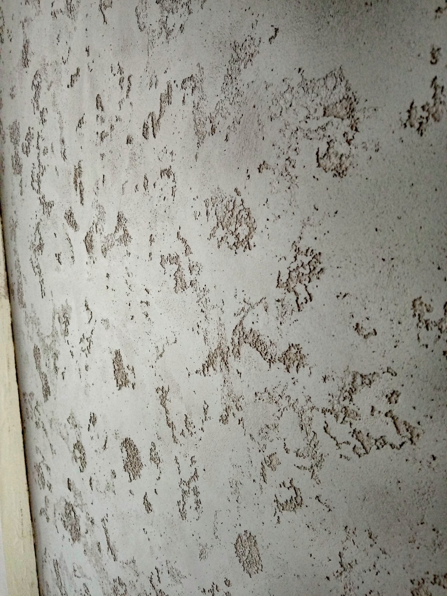 Szara ściana z wyraźną, nieregularną fakturą, przypominającą drobne, chropowate plamy i wgłębienia, oświetlona z boku, z fragmentem listwy przypodłogowej po lewej stronie.