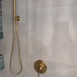 Złota deszczownica z termostatem zamontowana na szarych płytkach imitujących marmur. Widoczny fragment dekoracyjnej ściany z mozaiki.