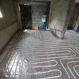 Instalacja ogrzewania podłogowego Roth na podkładzie z siatką w surowym wnętrzu, widoczne rury rozprowadzające i rozdzielacz z niebieskimi elementami.