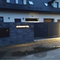 Nowoczesna, przesuwna brama wjazdowa z podświetleniem LED, zamontowana przy posesji z szarej kostki, z widoczną skrzynką gazową i podświetlonym adresem.