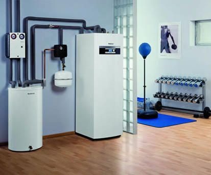 Biała instalacja grzewcza z dwoma zbiornikami, rurami i osprzętem w pomieszczeniu z parkietem, widoczne elementy wyposażenia siłowni w tle.