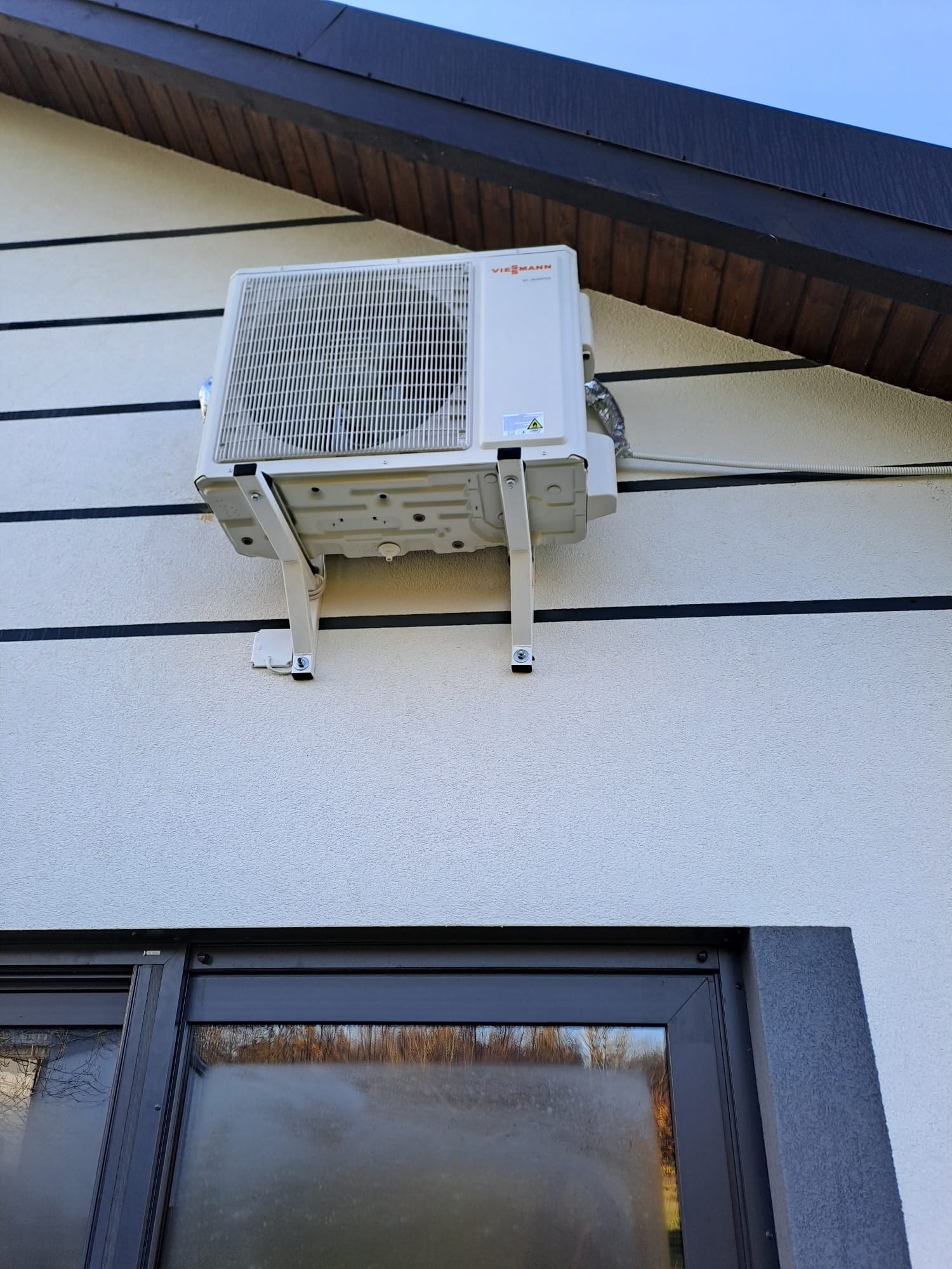 Biała jednostka zewnętrzna klimatyzacji marki Viessmann zamontowana na wspornikach na elewacji domu z widocznym oknem w dolnej części kadru.