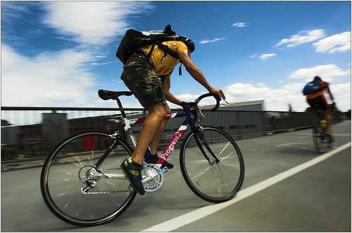 Kurier rowerowy w żółtej koszulce i z plecakiem dostarcza przesyłkę, jadąc po asfalcie w słoneczny dzień.