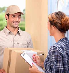 Kurier w beżowej czapce i koszuli wręcza paczkę kobiecie w kraciastej koszuli przy drzwiach. Kobieta trzymając paczkę, podpisuje dokument na podkładce.