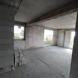 Surowy stan wnętrza domu w trakcie budowy, widoczne ściany z betonu komórkowego, surowy betonowy strop i wstawione okna z widokiem na zieleń na zewnątrz.