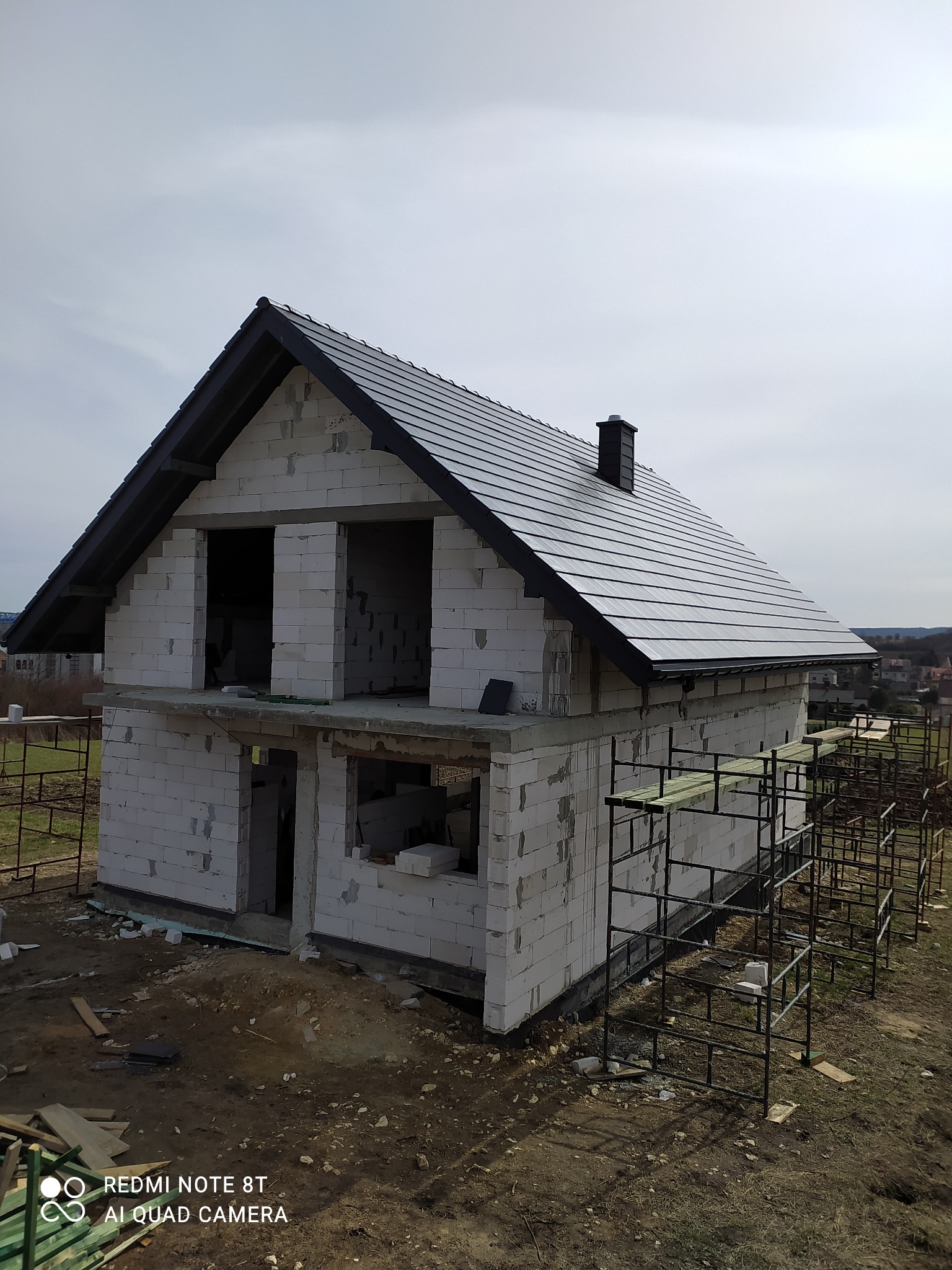 Surowy budynek z białych bloczków, z ciemną dachówką i kominem, widoczne rusztowania i elementy konstrukcyjne, na tle pochmurnego nieba i zabudowań w oddali.