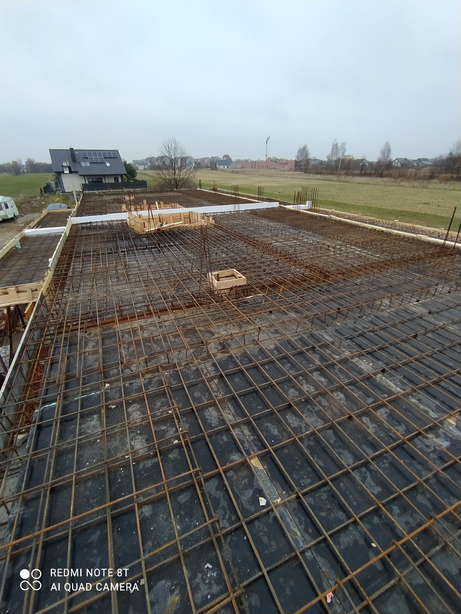 Zbrojenie płyty fundamentowej z widocznymi szalunkami i wystającymi prętami, w tle budynek mieszkalny z panelami słonecznymi i krajobraz.