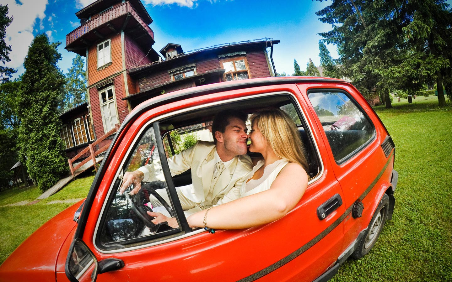 Para młoda w czerwonym Fiacie 126p, sfotografowana szerokokątnym obiektywem na tle drewnianego domu z wieżą, w słoneczny dzień.