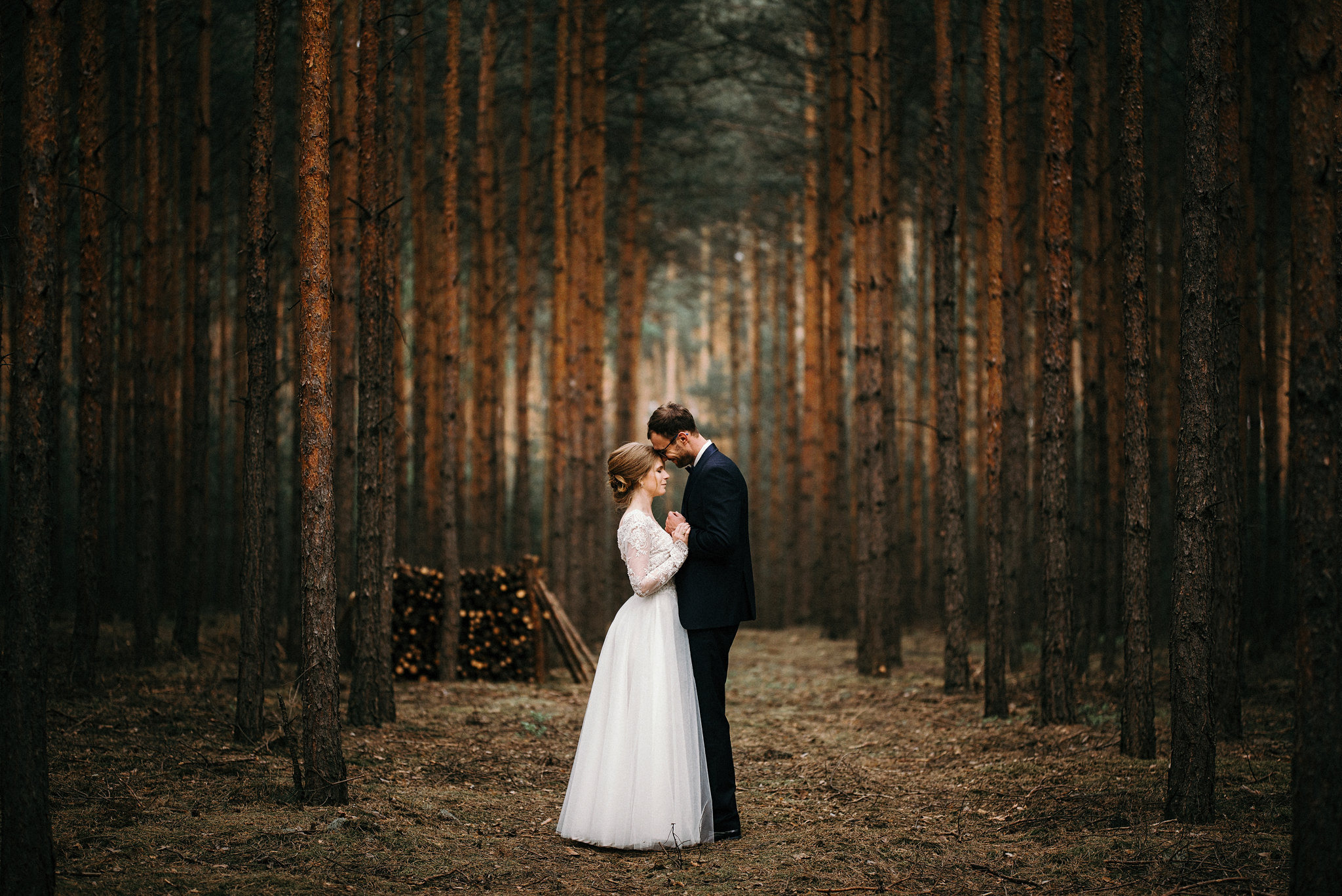Para młoda w lesie sosnowym: Panna młoda w koronkowej sukni z długim rękawem i pan młody w ciemnym garniturze, stojący blisko siebie na tle wysokich drzew. W tle stos drewna.
