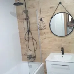 Nowoczesna łazienka z wanną obudowaną białą cegiełką, czarną armaturą, kabiną prysznicową z przezroczystego szkła, umywalką z szafką w kolorze białym oraz okrągłym lustrem zawieszonym na pasku.