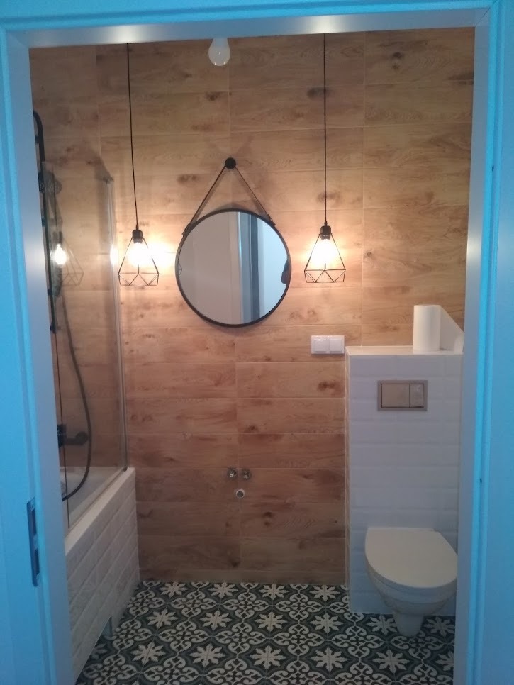 Nowoczesna łazienka z drewnopodobnymi płytkami ściennymi, okrągłym lustrem na pasku, wiszącymi lampami drucianymi, białą toaletą i wzorzystą podłogą w odcieniach zieleni i bieli, widok zza...