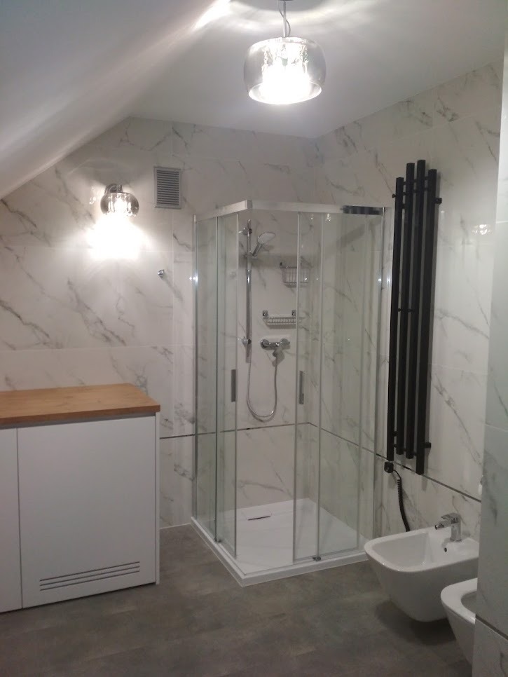 Nowoczesna łazienka na poddaszu z marmurowymi płytkami, kabiną prysznicową narożną, grzejnikiem drabinkowym i białą zabudową szafki z drewnianym blatem.