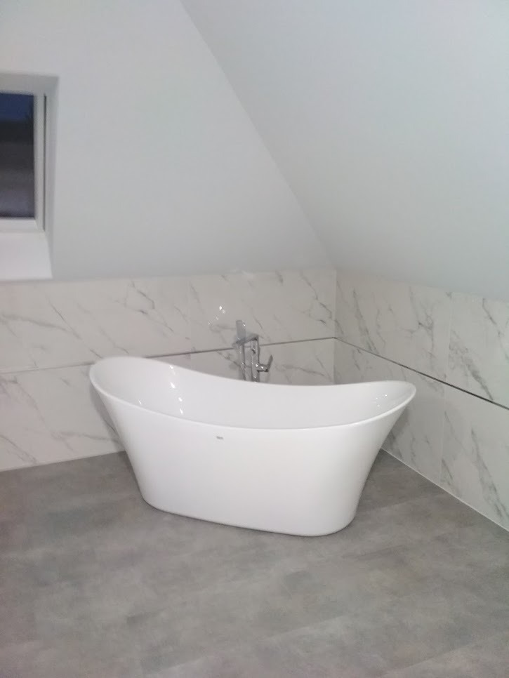 Wolnostojąca wanna w wykończonej łazience na poddaszu w Kielcach, z marmurowymi płytkami na ścianach i szarą podłogą.