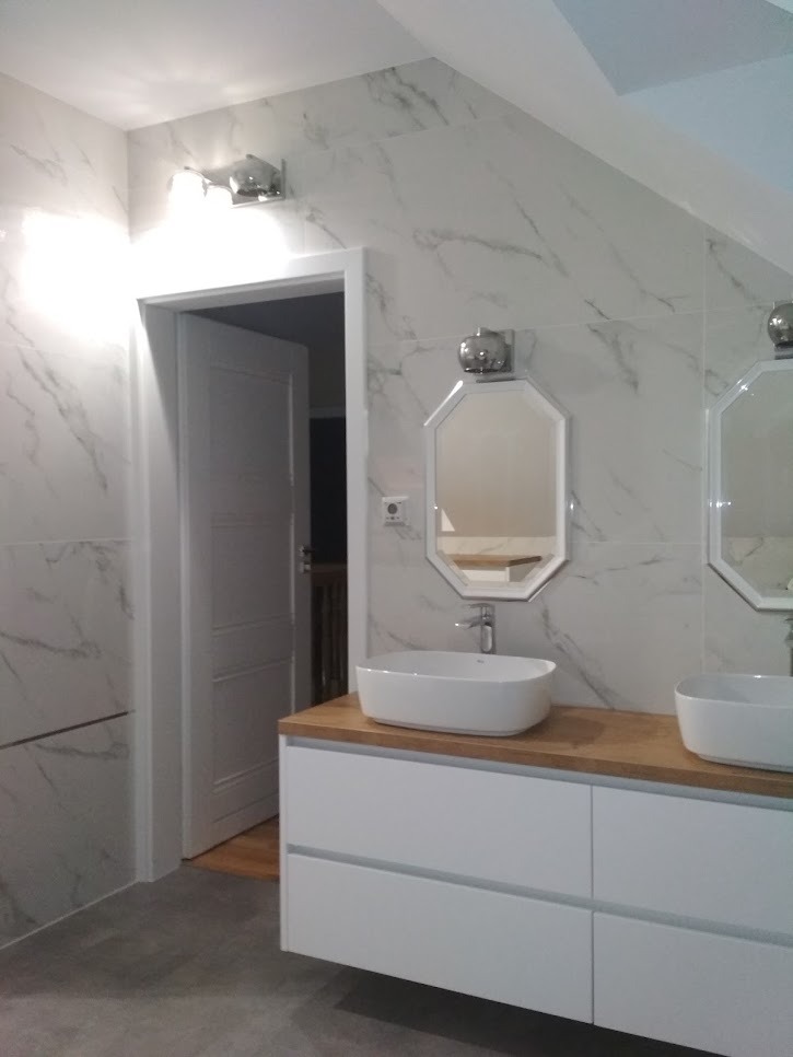 Nowoczesna łazienka z marmurowymi płytkami, dwiema owalnymi umywalkami na drewnianym blacie i białymi szafkami podwieszanymi, oświetlona chromowanymi lampami.