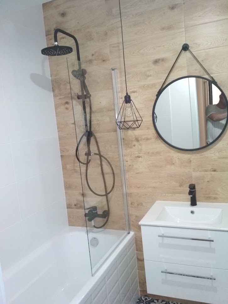 Nowoczesna łazienka z wanną obudowaną białą cegiełką, czarną armaturą, kabiną prysznicową z przezroczystego szkła, umywalką z szafką w kolorze białym oraz okrągłym lustrem zawieszonym na pasku.