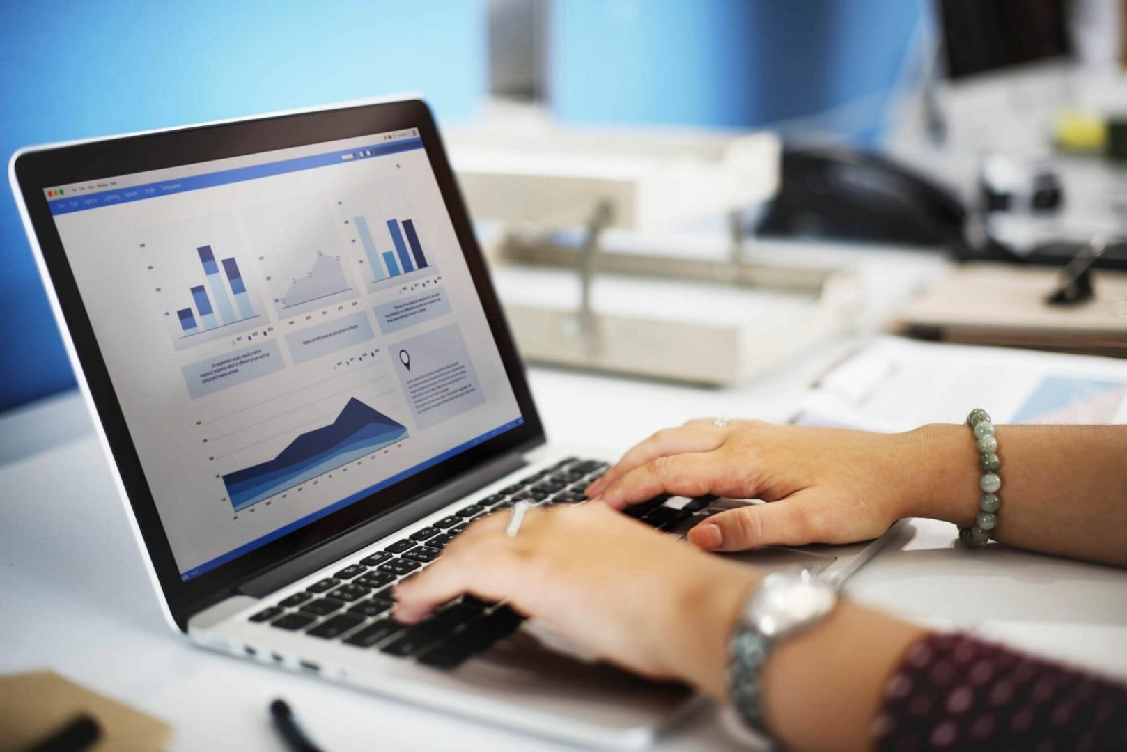 Osoba wpisuje dane do laptopa z otwartymi wykresami słupkowymi i liniowymi, widoczny zegarek na ręce i bransoletka z zielonych koralików.