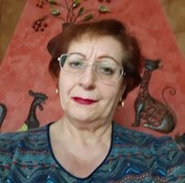 Portret kobiety w okularach, z krótkimi rudymi włosami i czerwonymi ustami, na tle ściany z dekoracjami w kształcie kotów.