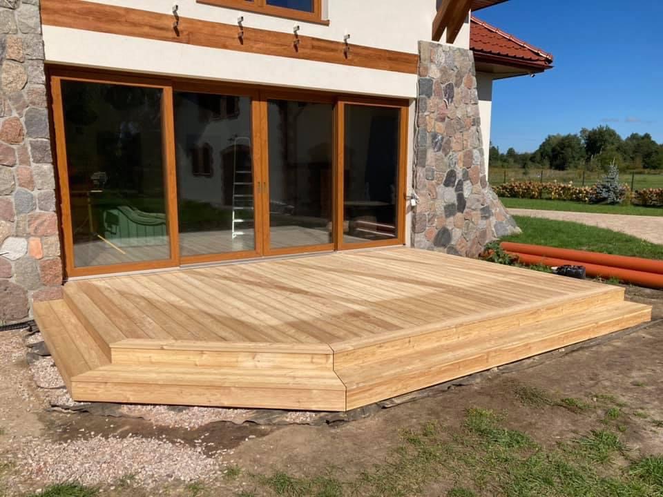 Jasny, drewniany taras z niskimi schodami w Piasecznie, przylegający do domu z dużymi przesuwnymi drzwiami i kamienną elewacją.
