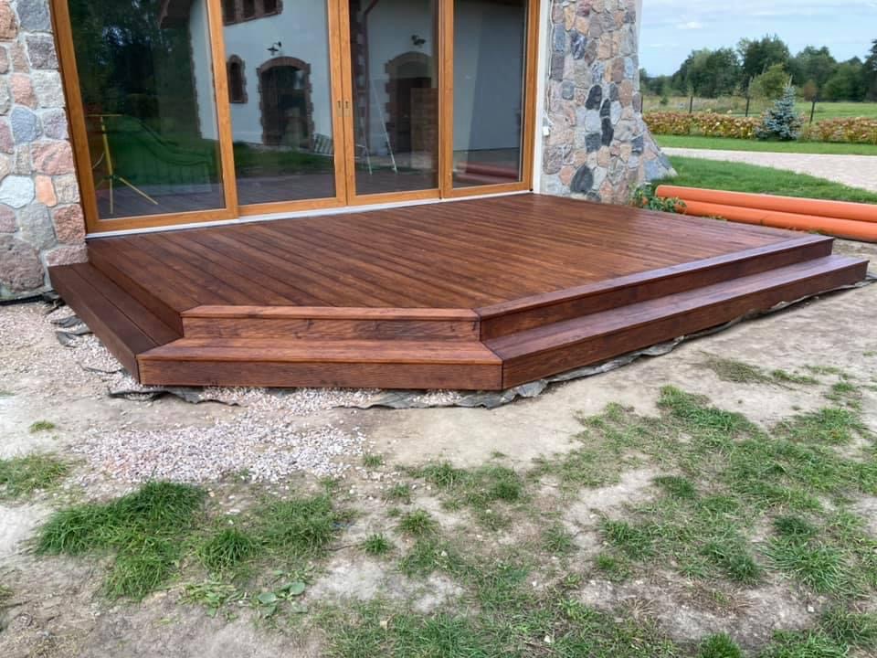 Nowoczesny taras z ciemnego drewna z dwoma stopniami, przylegający do kamiennego domu z dużymi oknami tarasowymi, widoczne pomarańczowe rury na trawniku.