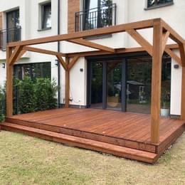 Drewniana pergola zadaszająca taras z desek w ciepłym odcieniu brązu, z dwoma stopniami wejściowymi, na tle białej elewacji domu z dużymi przesuwnymi drzwiami tarasowymi.