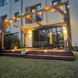 Drewniana pergola oświetlona girlandą żarówek, przylegająca do tarasu z desek, z widokiem na przesuwne drzwi balkonowe i elewację budynku.