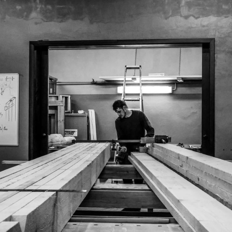 Czarno-białe zdjęcie stolarza pracującego w warsztacie nad obróbką drewnianych belek, widoczna drabina w tle i tablica z rysunkami technicznymi.