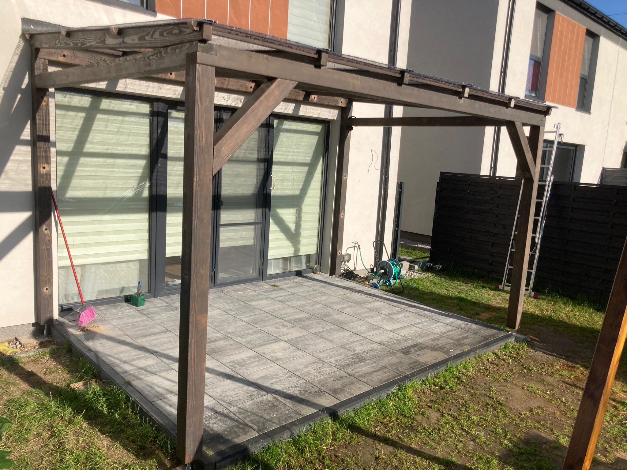 Nowoczesna pergola z ciemnego drewna, zadaszona, przylegająca do budynku z dużymi oknami tarasowymi, na wyłożonym kostką brukową tarasie, z widocznymi śladami niedawnych prac wykończeniowych...