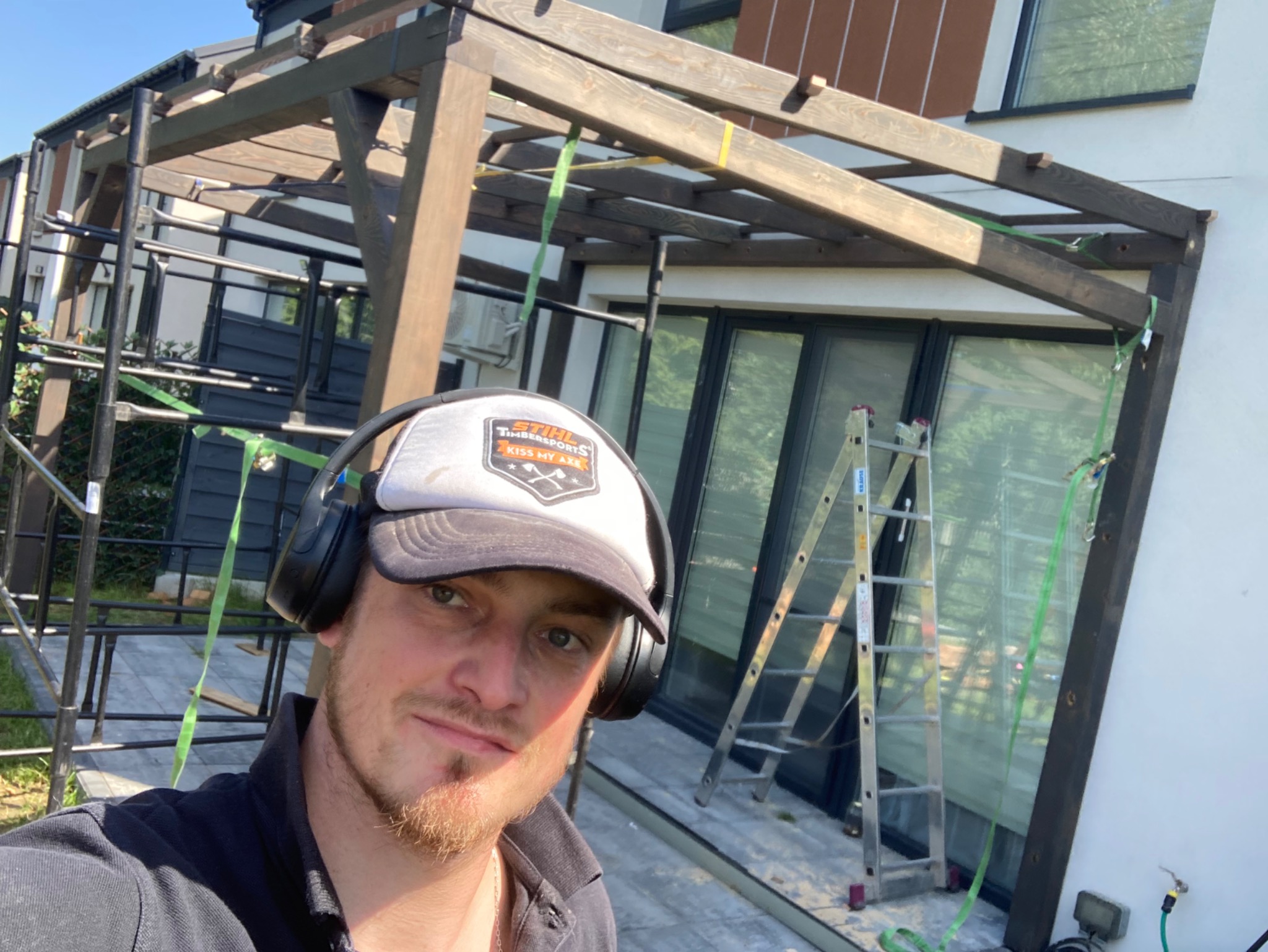 Mężczyzna w czapce i słuchawkach na tle konstrukcji drewnianej pergoli w trakcie montażu, widoczne rusztowanie i drabina oparta o okno.